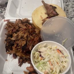 A foam takeout container holds pulled pork, a bun with meat, a cup of water, and a side of coleslaw, served for casual dining.