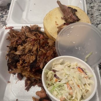 The Rub Bar-B-Que - A foam takeout container holds pulled pork, a bun with meat, a cup of water, and a side of coleslaw, served for casual dining. - Barbeque Near Me - Olathe, Kansas