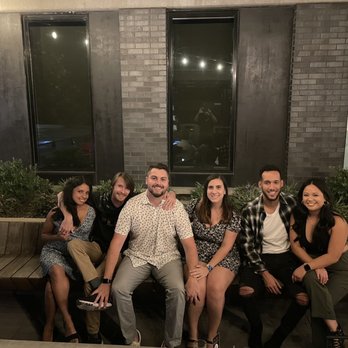 Gin Bar - Beautiful rooftop with great lighting and fire pits. Photo courtesy of our amazing bartender. - Six individuals are sitting on a curved wooden bench outdoors at night, with a dark brick building and large windows behind them. They are smiling and appear to be enjoying each other's company. The group includes three women and three men, dressed casually. The setting suggests a social gathering or night out. - Bars Near Me - Austin, Texas
