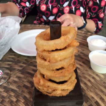 Clementines Too - 6 inch stack of onion rings. - A tall stack of fried onion rings is displayed on a small black stand on a wooden table, with dipping sauces and a drink nearby. - Seafood Near Me - Saint Joseph, Michigan