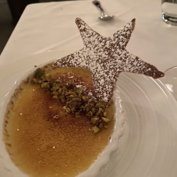 L'etoile Restaurant - Creme brûlée - A crème brûlée dessert is presented in a white ramekin with a caramelized sugar top. It is garnished with crushed nuts along one side and a star-shaped chocolate decoration dusted with powdered sugar, positioned upright. The dessert is on a white plate, with a spoon and a glass of water visible in the background, on a table set in a restaurant setting. - French Near Me - Edgartown, Massachusetts