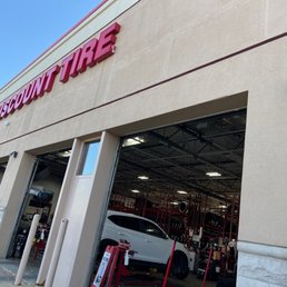 Junction road location. - The photo depicts the exterior of a Discount Tire store, with multiple open garage bays revealing the interior workspace and a vehicle inside. The store's sign is prominently displayed on the building facade. The area outside the store includes a sidewalk and parking space, with a clear blue sky overhead.