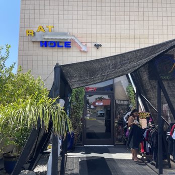 Rat Hole - Front of the store - A retail store with a neon sign reading 'RAT HOLE' on the exterior wall, featuring an outdoor clothing rack and a woman browsing items. - Vinyl Records Near Me - Riverside, California