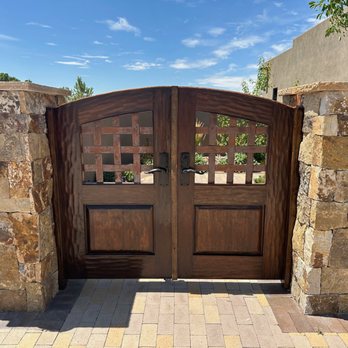 Authentic Wood Doors Carpentry - A double wooden gate with a curved top and metal handles is positioned between two stone pillars made of irregularly shaped stones. The gate features a lattice design at the top and solid panels at the bottom. The scene is outdoors with a paved walkway in front, and the background includes trees and a building wall under a partly cloudy blue sky. - Carpenters Near Me - Santa Fe, New Mexico