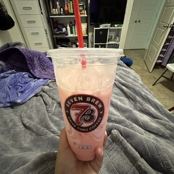7 Brew Coffee - Strawberry , Peach, Mango Fiiz w/ cream - A plastic cup with a Seven Brew Coffee logo contains a pink, fruit-flavored beverage with a red straw, held over a bed with a gray blanket. - Coffee & Tea Near Me - Rio Rancho, New Mexico