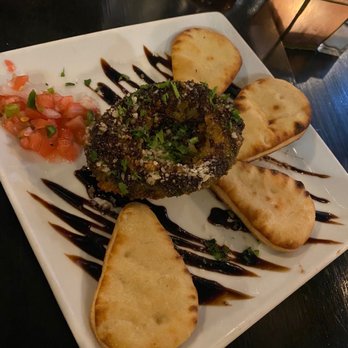 Debajo Tapas ABQ - Aguacate Frito - A square white plate holds fried avocado slices arranged around a central breaded and fried avocado half topped with herbs. The plate is garnished with a drizzle of balsamic glaze and a small portion of tomato salsa. The setting includes drinks and a candle in the background, indicating a dining environment. - Spanish Near Me - Albuquerque, New Mexico