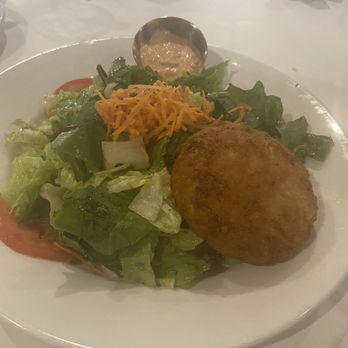 Cafe Lynn - Crabcake salad - A salad with mixed greens, shredded cheese, and a fried crab cake on a white plate, accompanied by a small cup of dressing. - Seafood Near Me - Mandeville, Louisiana
