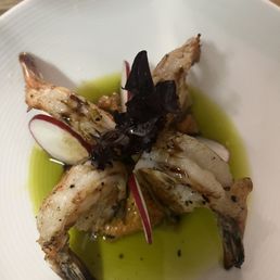 The most delicious shrimp appetizer! Rich in flavor and texture! - A seafood dish with shrimp arranged around a central garnish of black leaves or herbs, garnished with thin radish slices. The dish is served with a green oil or sauce pooled at the bottom of a white, textured plate. The presentation emphasizes fresh ingredients and a minimalist style.