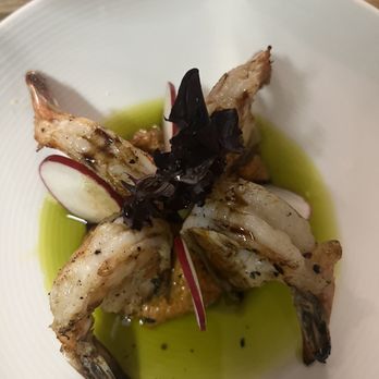 Caffeine's Midtown - The most delicious shrimp appetizer! Rich in flavor and texture! - A seafood dish with shrimp arranged around a central garnish of black leaves or herbs, garnished with thin radish slices. The dish is served with a green oil or sauce pooled at the bottom of a white, textured plate. The presentation emphasizes fresh ingredients and a minimalist style. - American Near Me - West Springfield, Massachusetts