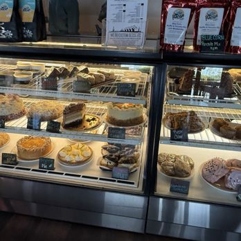 Range Cafe - Sweets 10% off of you order dessert before your meal. - A refrigerated display case contains a variety of desserts including cakes, cookies, and pastries. The top shelf features slices of cake, while the lower shelves hold cookies and other baked goods. Small labels identify the desserts, such as carrot cake, pie, and banana pudding. The case is illuminated, and the background shows a counter with coffee bags and a menu sign. - Breakfast & Brunch Near Me - Albuquerque, New Mexico