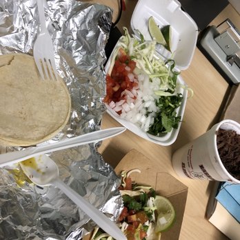 Barbacoa El Primo - Beef barbacoa - The image shows a taco with toppings like cilantro, onions, and salsa, along with a styrofoam container of chopped vegetables and a cup of shredded beef on a wooden table. - Mexican Near Me - Albuquerque, New Mexico