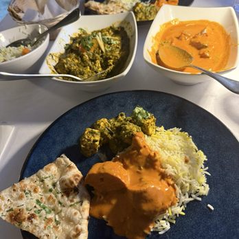 Mint - Indian Lakeside Dining - The image shows a blue plate containing rice, naan bread, and a curry dish with orange sauce. In the background, there are two bowls of Indian dishes, one with a greenish curry and the other with an orange curry, along with a person eating at the table. The setting appears to be a restaurant with a white tablecloth. - Indian Near Me - Lee, Massachusetts