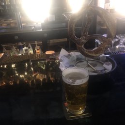 Goose Island And Jumbo Bavarian Pretzel - A bar counter scene includes a large pretzel on a metal stand, placed on a tray with paper wrapping, and a glass of beer with bubbles, sitting on a coaster. The background shows bar equipment, bottles, and glasses, with bright light sources creating glare and reflections on the surfaces.
