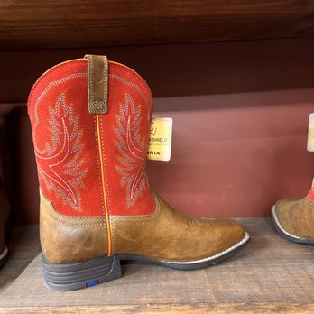 Tecovas - A pair of cowboy boots with red leather shafts featuring embroidered leaf-like patterns and brown leather foot sections are shown on a wooden store shelf. The boots have a zipper on the side and a low heel, with tags indicating they are for sale. The background includes other footwear and a red wall. - Leather Goods Near Me - Austin, Texas