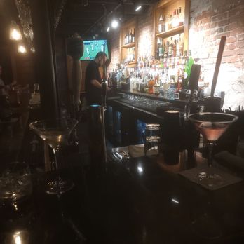 1883 Lounge - 1883 Lounge - The image depicts a bar with a bartender preparing drinks behind a counter. Shelves filled with various liquor bottles are mounted on a brick wall, illuminated by focused lighting. A television mounted on the wall shows a sports game. The foreground includes glasses and bar equipment, with dim ambient lighting creating a cozy atmosphere. - American Near Me - Onset, Massachusetts