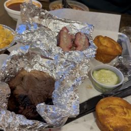 Rib tips, brisket, and cornbread - The image shows a person dining at a restaurant with a spread of barbecue dishes. The person is holding a fork and wearing a dark jacket with a small logo. The table features foil-wrapped ribs, cornbread muffins, a bowl of soup, and small containers of sauces and sides. The setting appears casual, with a brick wall in the background and a variety of food items typical of barbecue cuisine.