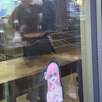 Dragon Sushi - Wait staff with butt on table and smoking rather than waiting tables - The image shows several individuals inside a commercial establishment, possibly a restaurant or shop, viewed through a glass window. Reflections on the glass partially obscure the scene, which includes a wooden counter and a cartoon character sticker with pink hair and glasses attached to the glass. The interior features colorful signage and displays, indicating a lively environment. - Sushi Bars Near Me - Albuquerque, New Mexico