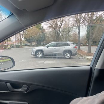 Costco Wholesale - The security guard baby sitting us. - The image shows a silver SUV parked in an outdoor parking lot, with trees and a sidewalk visible in the background. The photo is taken from inside a vehicle, capturing part of the dashboard, door, and a person's arm. The parking lot appears mostly empty, with clear weather and leafless trees indicating a possible fall or winter season. - Wholesale Stores Near Me - Eugene, Oregon