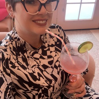 Muerta Lashes - Tooth gem - A woman with short, dark hair and large, stylish glasses is smiling at the camera. She is wearing a black and white patterned outfit and earrings. She holds a large glass of pink cocktail garnished with a lime slice and a pink straw. The background shows a door with glass panes and a tiled floor, indicating an indoor setting. - Eyelash Service Near Me - Gilbert, Arizona
