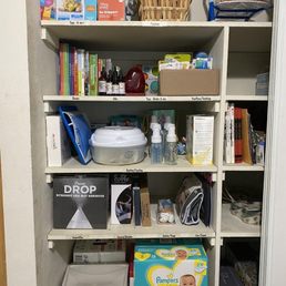 After - The image shows a white, five-shelf storage unit filled with a variety of household and baby care items. The top shelf holds toys, a basket, and a small blue stroller. The second shelf contains books, oils, and a box of toiletries. The third shelf has bottles of disinfectant spray, a container, and a box of medical supplies. The fourth shelf features an ultrasonic cool mist humidifier, a cover, and a bag. The bottom shelf holds boxes of Pampers diapers and a trash bin. The shelves are neatly organized with labeled sections for different categories of items.