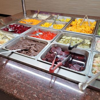 Amherst Buffet - A buffet setup at Amherst Buffet includes multiple metal trays filled with fruits, desserts, and cold cuts. Visible items include sliced oranges, cherries, jellies, and sliced cucumbers, with serving utensils in each tray. The buffet is arranged on a polished granite counter with a glass sneeze guard above. The presentation is organized and clean, offering a range of options for diners. - Chinese Near Me - Tonawanda, New York
