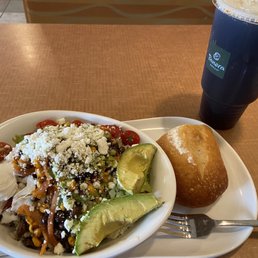 A meal is presented on a white tray with a bowl of salad containing cherry tomatoes, avocado slices, shredded cheese, and a poached egg. A bread roll is placed beside the salad, and a large dark soda in a plastic cup with a lid and straw is positioned behind the tray. The setting appears to be in a casual dining area, likely a cafe or fast-food restaurant.