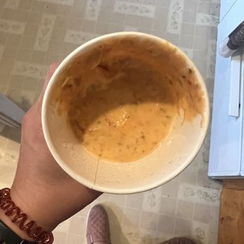 Francisca's - A person is holding a paper cup containing leftover sauce or soup, with visible herbs and liquid residue inside. The photo is taken from a top-down perspective, showing the person's hand, wristwatch, and bracelet. In the background, a tiled floor, a person wearing flip-flops, and part of a kitchen or dining area are visible, indicating an indoor setting. - Mexican Near Me - Farmington, New Mexico