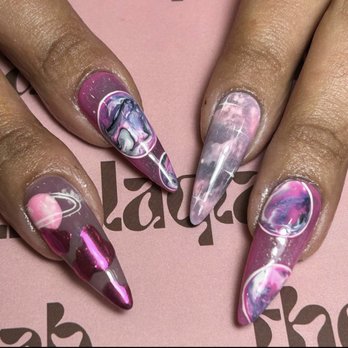Thelaqlab - The image shows four long, stiletto-shaped nails with intricate galaxy-inspired marble nail art. The nails display swirling patterns of pink, purple, white, and black, resembling cosmic scenes. The nails are glossy and well-manicured, with each nail having a unique design. The background is pink with partially visible text, emphasizing the detailed nail art. - Nail Salons Near Me - Brooklyn, New York