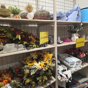 Beall's Outlet Store - Metal shelving units hold various decorative items such as artificial pumpkins, floral arrangements, and holiday-themed decor. Some items are marked with yellow clearance signs. The shelves also contain other household items like blankets, vases, and storage containers, arranged in a retail setting. - Outlet Stores Near Me - Buckeye, Arizona