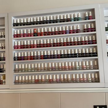 Strong Nail City - Colors! - A display of numerous nail polish bottles arranged on white shelves in a salon or beauty store. The bottles are organized by color, including shades of pink, red, purple, green, and neutral tones. The shelves are mounted on a wall, with a tiled surface below. The image captures the variety of nail polish options available for customers. - Nail Salons Near Me - Winnetka, Illinois