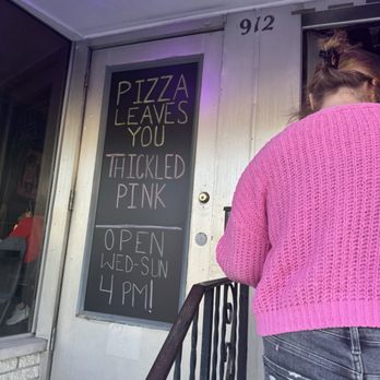 Molten Monkey - Outside - A woman wearing a pink knitted sweater and jeans is standing outside a pizza restaurant, next to a chalkboard sign with the shop's hours and a humorous message. - Pizza Near Me - Madison, Wisconsin