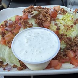 Wedge salad - A wedge salad served on a white rectangular plate includes iceberg lettuce cut into wedges, topped with chopped tomatoes, onions, and seasoned ground beef. A small cup of ranch dressing is placed in the center of the plate. The background shows a dark table surface with a glass of water and a small container of pepper.