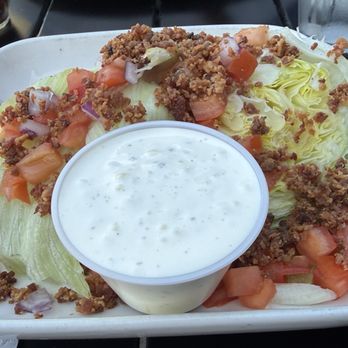 Treehouse Pub & Eatery - Wedge salad - A wedge salad served on a white rectangular plate includes iceberg lettuce cut into wedges, topped with chopped tomatoes, onions, and seasoned ground beef. A small cup of ranch dressing is placed in the center of the plate. The background shows a dark table surface with a glass of water and a small container of pepper. - Pizza Near Me - Bettendorf, Iowa