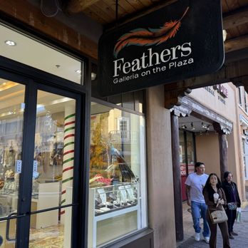 Feathers Galleria - The image depicts the storefront of Feathers Galleria on the Plaza, with a black sign featuring a feather graphic and the store name. The storefront has large glass windows displaying jewelry and decorative items inside. Pedestrians are walking along the sidewalk, and neighboring shops, including Häagen-Dazs, are visible in the background. The scene is set during daylight hours with clear weather. - Jewelry Near Me - Santa Fe, New Mexico