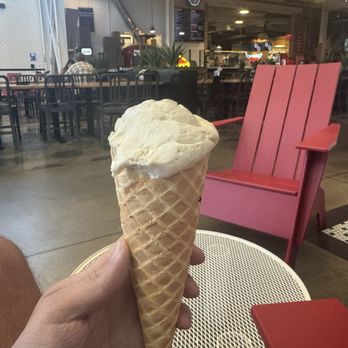 Moo Thru - A hand holds a vanilla ice cream cone in a cafe setting with pink chairs and tables, and a menu board in the background. - Ice Cream & Frozen Yogurt Near Me - Charlottesville, Virginia