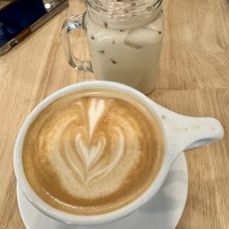 Oat milk latte - A latte with foam art in a white ceramic cup and saucer is placed on a wooden table. Behind it, there is a mason jar filled with iced coffee, milk, and ice cubes, topped with a splash of liquid. A smartphone and a utensil are also visible on the table.