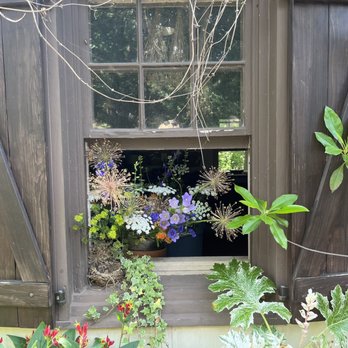 Chanticleer Foundation - A window with dark wooden shutters, one of which is slightly open, shows a flower box filled with a variety of colorful flowers and green plants. The window is part of a wooden structure, and some plants and vines are visible around it, adding to the garden-like setting. - Botanical Gardens Near Me - Wayne, Pennsylvania