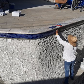 Baccaro Pools - A worker, dressed in a white long-sleeve shirt, blue jeans, and a straw hat, is standing at the edge of a swimming pool, reaching up to work on the pool's border. The pool is partially constructed, with textured concrete walls and a tiled coping. Nearby, there is a small table with a bucket, and the surrounding area has construction tools and materials, indicating ongoing pool construction or renovation. - Pool Cleaners Near Me - Walnut, California