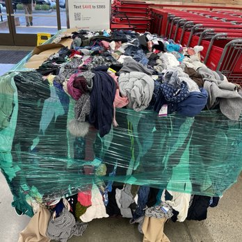 Savers - How clothes are brought in...interesting. - A large, wrapped bundle of used clothing is displayed on the floor of a thrift store, with various garments including sweaters, shirts, and scarves visible. The pile is secured with plastic wrap, and shopping carts are lined up behind it. The store entrance and a sign promoting text deals are also visible in the background. - Thrift Stores Near Me - Walnut Creek, California