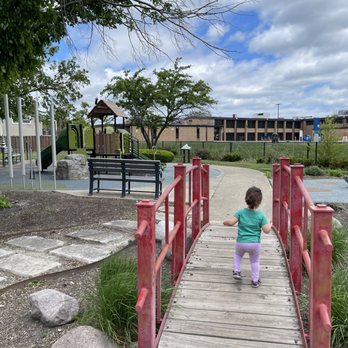 Overhill Park - A young child with curly hair, wearing a turquoise shirt and purple pants, is walking across a small wooden bridge with red railings in a playground. The playground features a slide, benches, and a turtle sculpture, with trees and a building in the background under a partly cloudy sky. - Playgrounds Near Me - Morton Grove, Illinois
