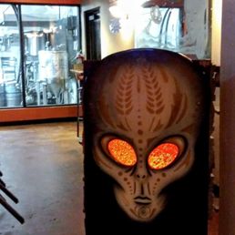 An alien-themed sign with glowing orange eyes is displayed inside a brewpub, featuring graffiti-style artwork and a reflective surface.