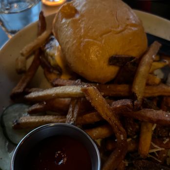 The Sample Room - Bottoneau burger - A burger with a bun, fries, and a small cup of dipping sauce on a plate. The burger appears to have cheese and possibly other toppings. The fries are scattered around the plate, and there are slices of cucumber and a pickle on the side. The background includes glasses of water and a lit candle, suggesting a dining setting. - New American Near Me - Minneapolis, Minnesota