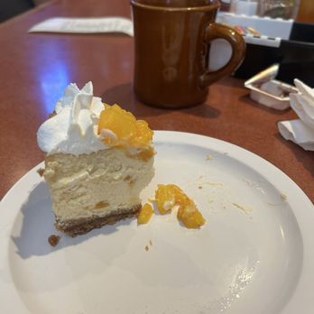 Old School Grub - Peach Cheesecake - A partially eaten slice of peach cheesecake topped with whipped cream and peach pieces is placed on a white plate. The cheesecake has a graham cracker crust and a creamy filling. In the background, there is a brown ceramic coffee mug, along with condiments and utensils on a table in a restaurant setting. - Burgers Near Me - Mesa, Arizona