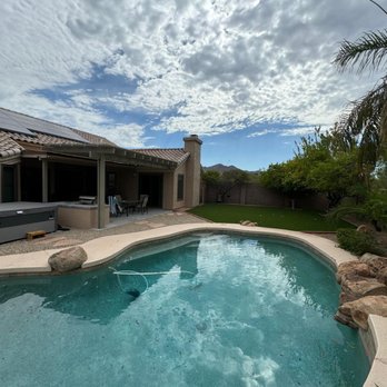 E.L. Construction - After new patio, outdoor kitchen, pergola - The image shows a backyard with a swimming pool that has a curved shape, surrounded by a concrete deck. Adjacent to the pool is a patio area with outdoor furniture, including a table and chairs. The yard includes landscaped elements such as trees, rocks, and a grassy area, with a house in the background that has solar panels on the roof. The sky is partly cloudy with patches of blue visible. - General Contractors Near Me - Phoenix, Arizona