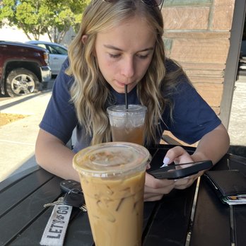 CB Kitchen - Cold brew - A woman with blonde wavy hair and sunglasses on her head is sitting at an outdoor cafe table, drinking iced coffee through a straw while looking at her phone. - Burgers Near Me - Willows, California