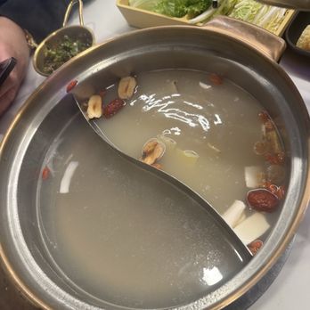 Coco Hotpot - 2 soup combo- coconut and healthy herbal chicken - A stainless steel hotpot with a divider in the middle, containing two different broths: one milky with goji berries and herbs, and the other clear. The hotpot is placed on a table with a tray of fresh vegetables, including lettuce and leafy greens, and other dining items. The setting suggests a communal dining experience, likely at a restaurant specializing in hotpot cuisine. - Hot Pot Near Me - Flushing, New York