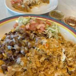 Burrito bowl and chicken tamale - A plate of Mexican cuisine featuring rice, refried beans topped with cheese, shredded lettuce, diced tomatoes, and two small containers of salsa, with a second plate of food in the background.