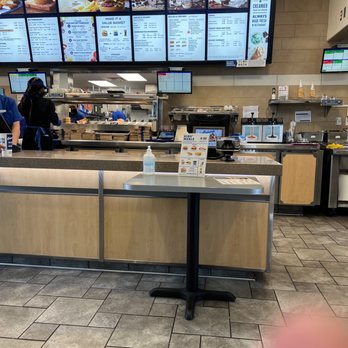 Culver's - Clean and efficient location - The image shows the interior of Culver's restaurant, featuring a counter area with digital menu screens displaying various meal options, including burgers, desserts, and value meals. Staff members are visible behind the counter, preparing food and wearing uniforms. A small table with a bottle of hand sanitizer and a promotional sign for kids' meals is positioned in front of the counter. The setting includes tiled flooring and a neutral wall finish, with additional screens and equipment visible in the background. - Burgers Near Me - Gilbert, Arizona