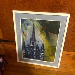 A framed watercolor painting depicts a castle with multiple spires and towers, rendered in shades of blue and black. The artwork is surrounded by a light blue mat and is leaning against a wooden wall on a wooden floor. The painting features abstract elements and swirling patterns in the sky area.