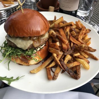 501 Local - Angus burger - A burger with a shiny, toasted bun contains melted cheese, caramelized onions, and fresh greens, served alongside a generous portion of French fries on a white plate. The setting includes a glass of red wine, a bottle of wine, and a potted plant in the background, indicating an outdoor dining environment at a restaurant. - New American Near Me - Winnetka, Illinois